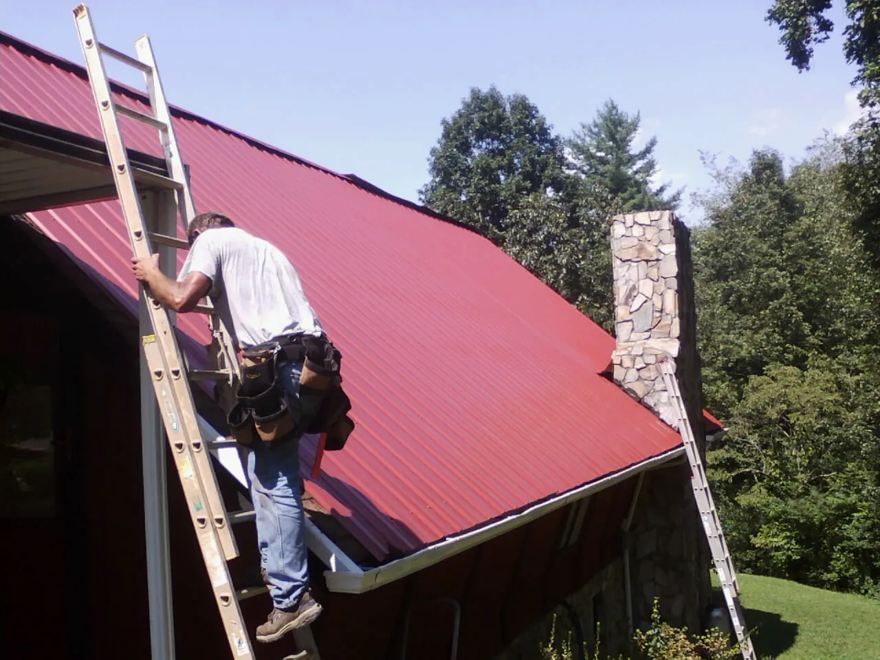 Roofer on a ladder beside a red metal roof and stone chimney in Vernonia